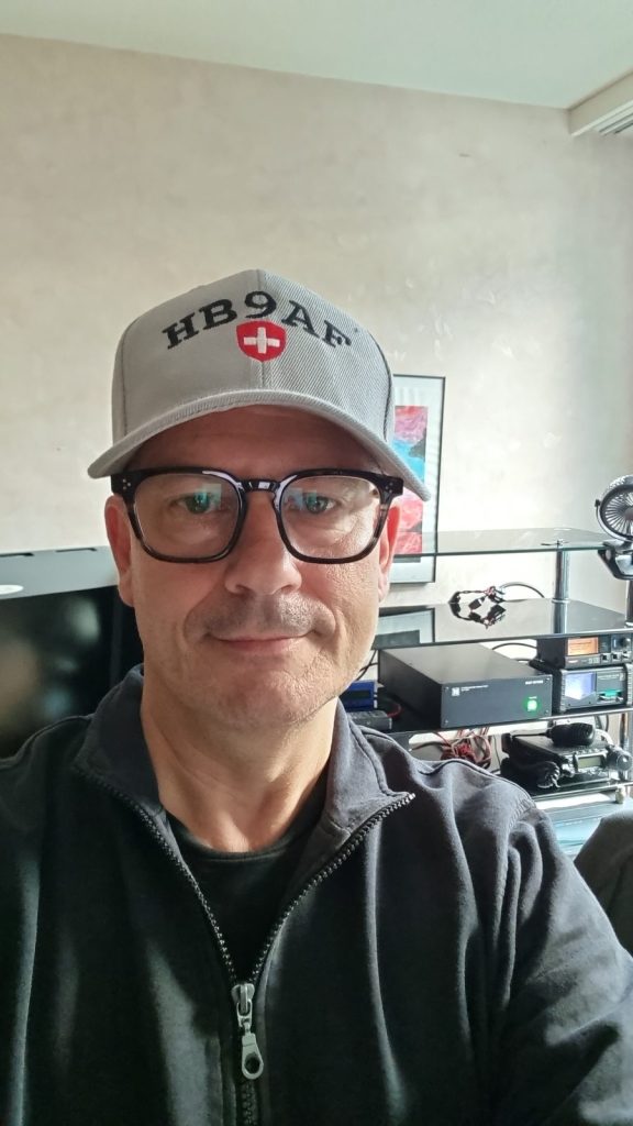 Chris HB9HJI mit HB9AF-Cap im Shack, im Hintergrund die Station
